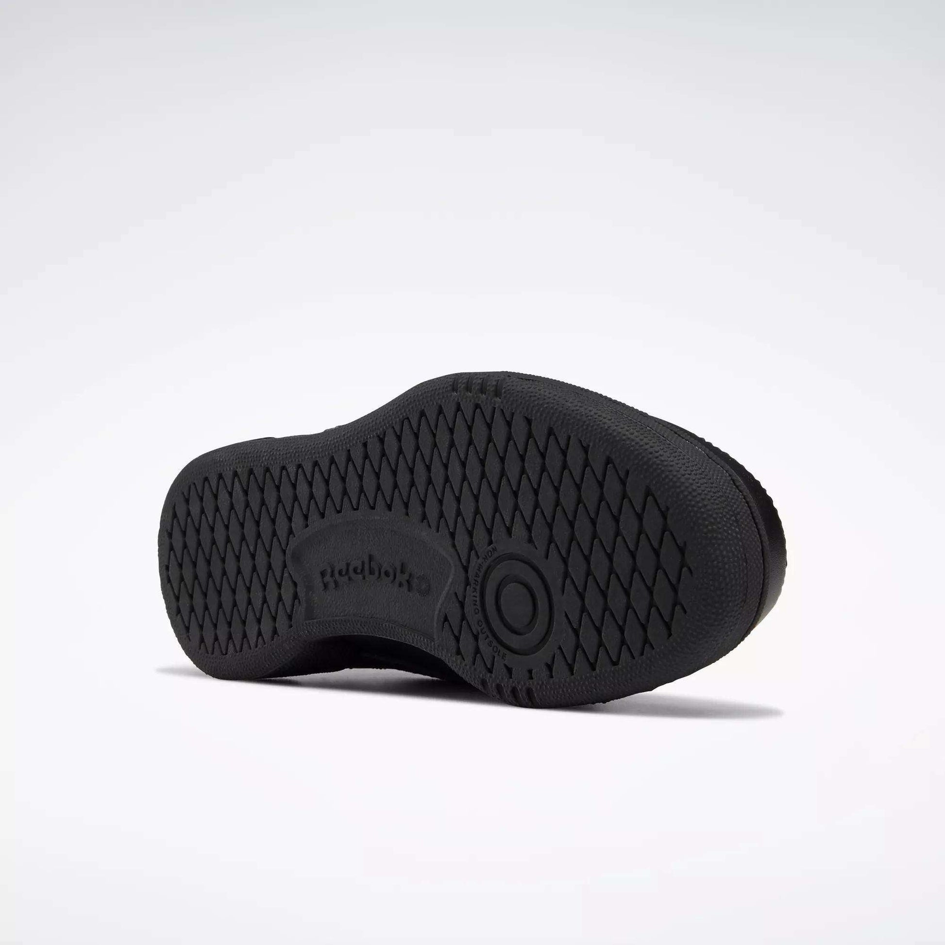 The image shows the sole of a black Reebok Kids' Club C Shoe - Grade School, with a high-abrasion rubber outsole, diamond-patterned grip, Reebok logo, and a circular design at the center. The background is plain white.
