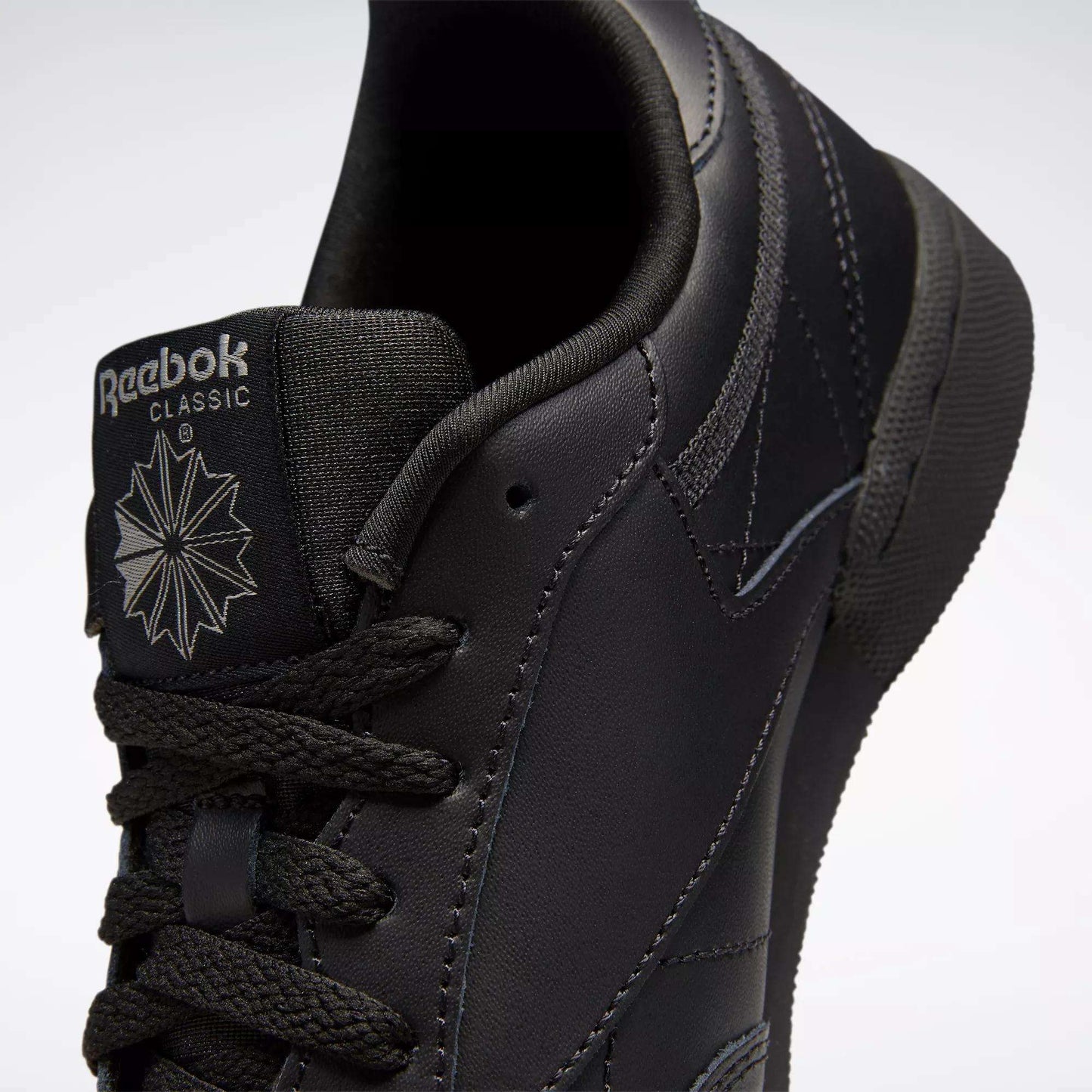 Close-up of black Reebok Kids' Club C Shoes (Grade School), featuring smooth leather uppers, the tongue with Reebok logo and starburst design, black laces, and detailed stitching.
