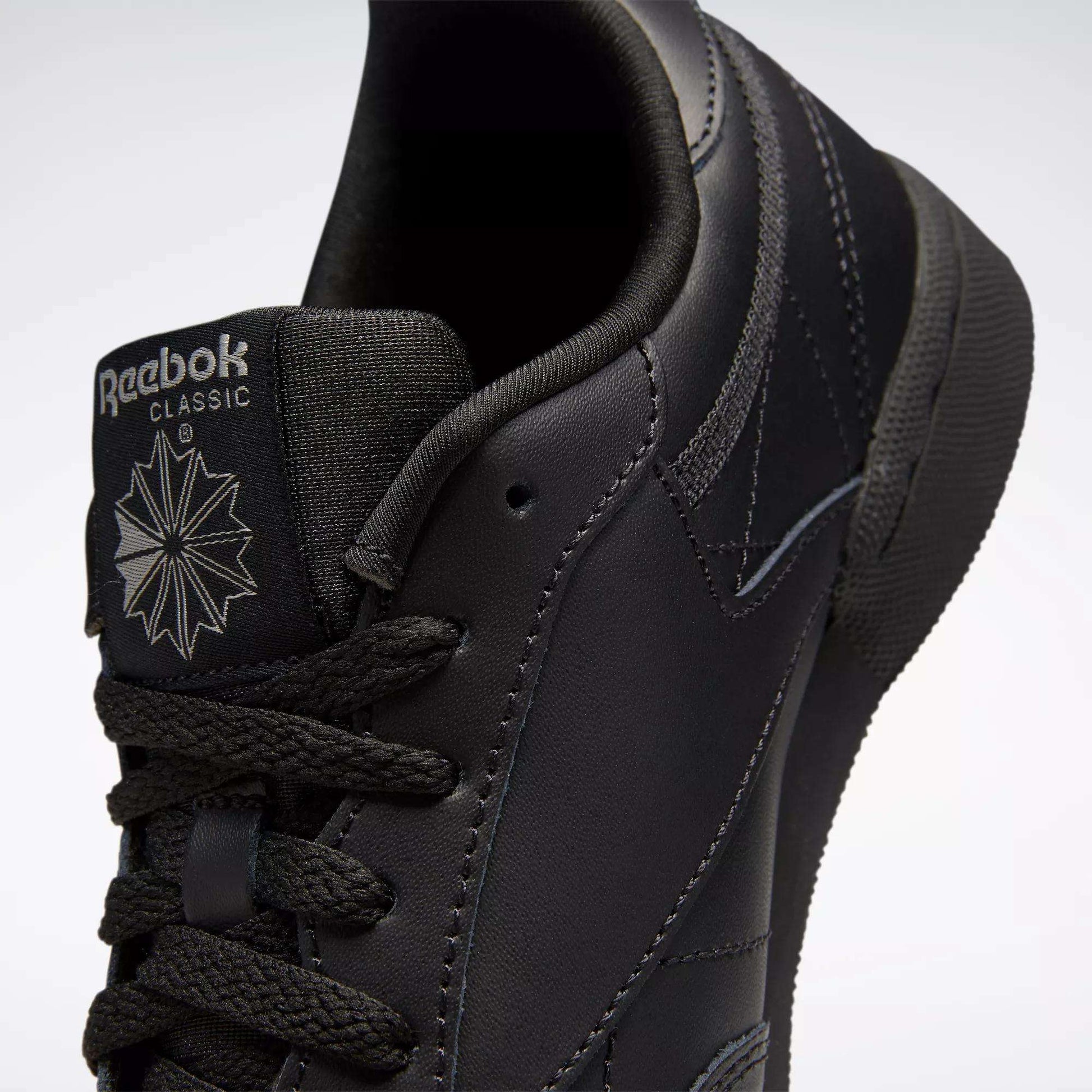 Close-up of black Reebok Kids' Club C Shoes (Grade School), featuring smooth leather uppers, the tongue with Reebok logo and starburst design, black laces, and detailed stitching.