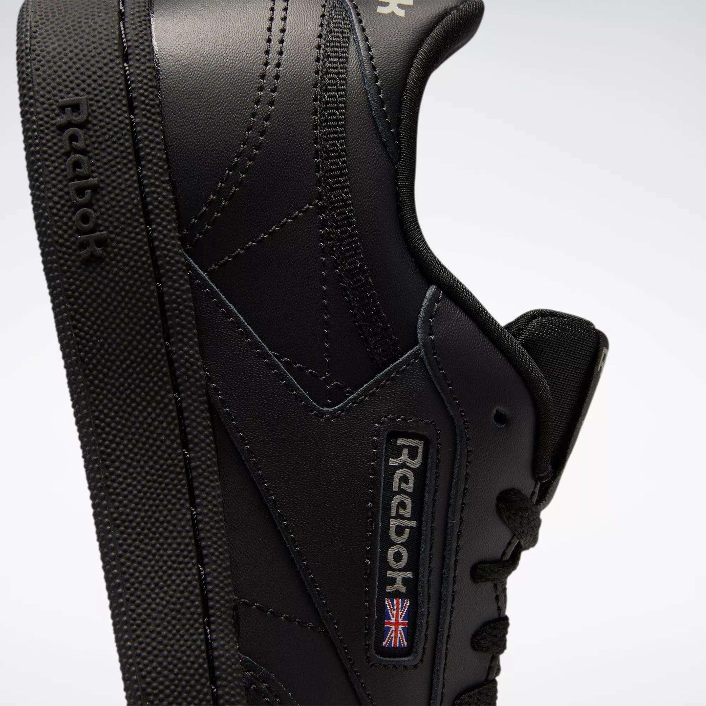 Close-up of Reebok Kids' Club C Shoes - Grade School in black, featuring a side view with visible stitching, soft leather uppers, textured sole, and Reebok branding with a small Union Jack flag.