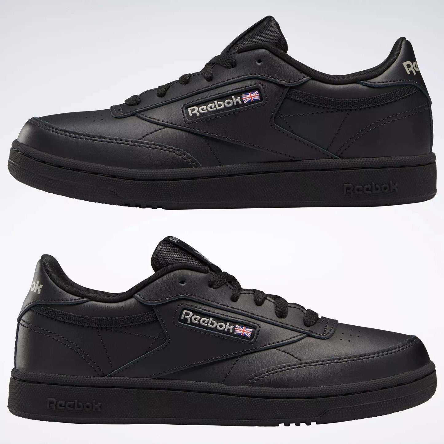 Two black Reebok Kids' Club C Shoes - Grade School are shown from the side, featuring soft leather uppers, low-top design, black soles, and the Reebok logo with a small British flag. The classic court shoe stands against a light background.