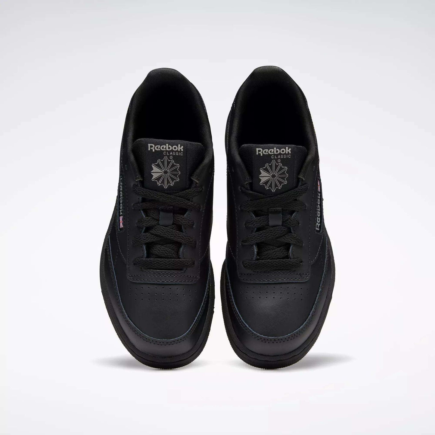 A pair of Kids' Club C Shoes - Grade School by Reebok, in black leather with perforated toe details, are shown from above. The classic court design features black laces and logos, positioned side by side on a plain light background.
