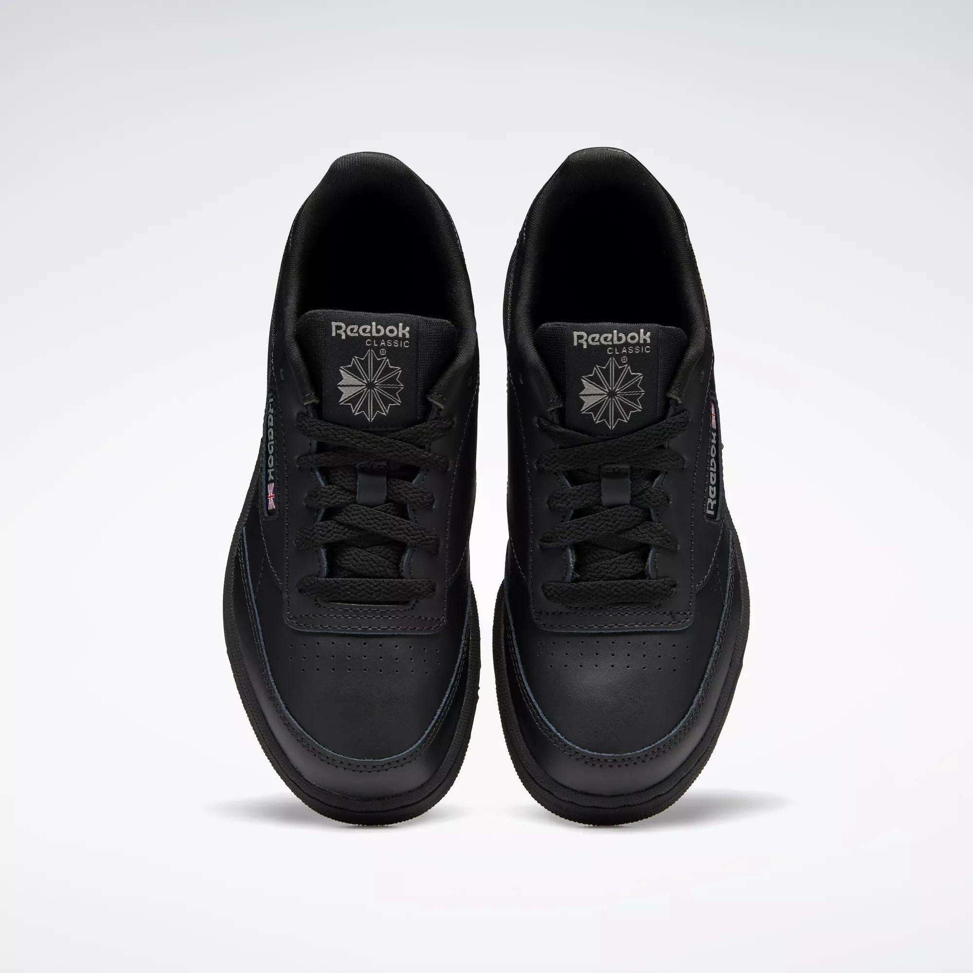 A pair of Kids' Club C Shoes - Grade School by Reebok, in black leather with perforated toe details, are shown from above. The classic court design features black laces and logos, positioned side by side on a plain light background.