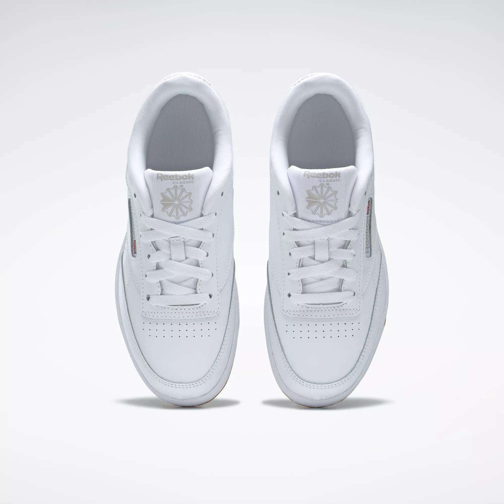 A pair of Kids' Club C Shoes - Grade School by Reebok, seen from above, with clean white leather uppers, laces, perforated toes, and Reebok logos on the tongues. The classic court style is displayed side by side on a light background.