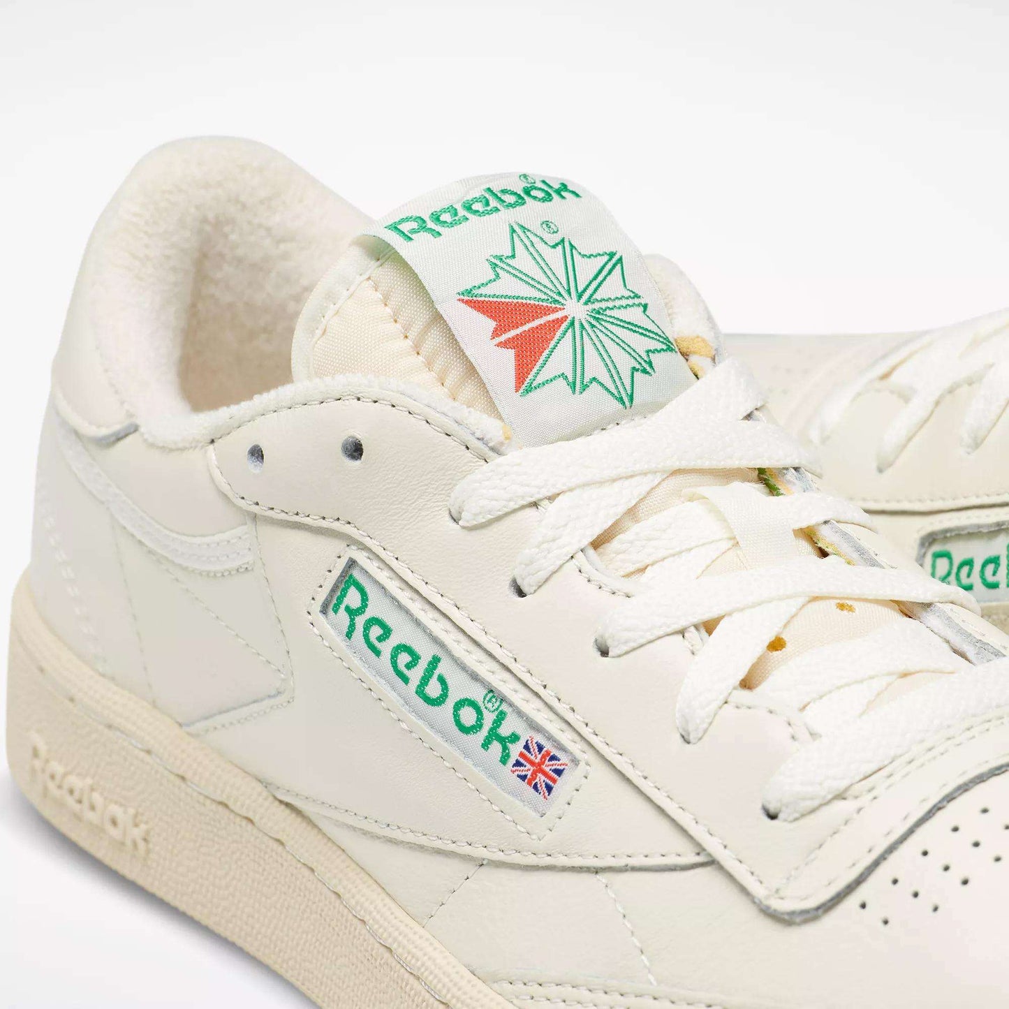 Close-up of off-white Reebok Club C 85 Vintage Shoes with green logos, white laces, and the British flag on the side. The tongue shows a green and red Reebok graphic, and the leather upper provides lightweight cushioning.