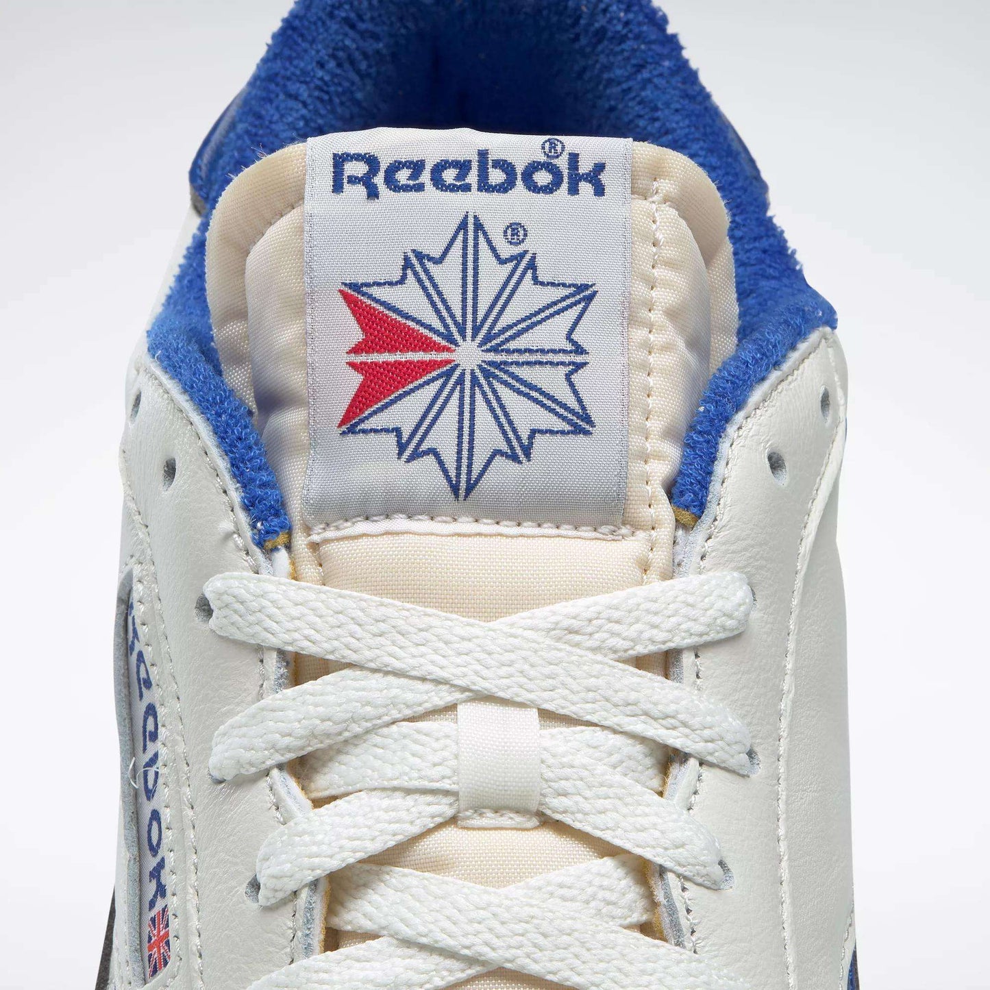 Close-up of Reebok Men's Club C Revenge Vintage Shoes, showing the white leather, blue logo on the tongue, red and blue starburst graphic, vintage-style laces, and blue collar lining for classic retro appeal.