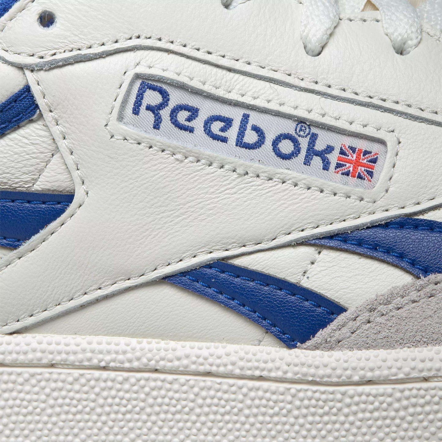 Close-up of Reebok Men's Club C Revenge Vintage Shoes in white leather with blue accents. The side features the Reebok logo and British flag above a textured white sole, showcasing its retro tennis-inspired design.