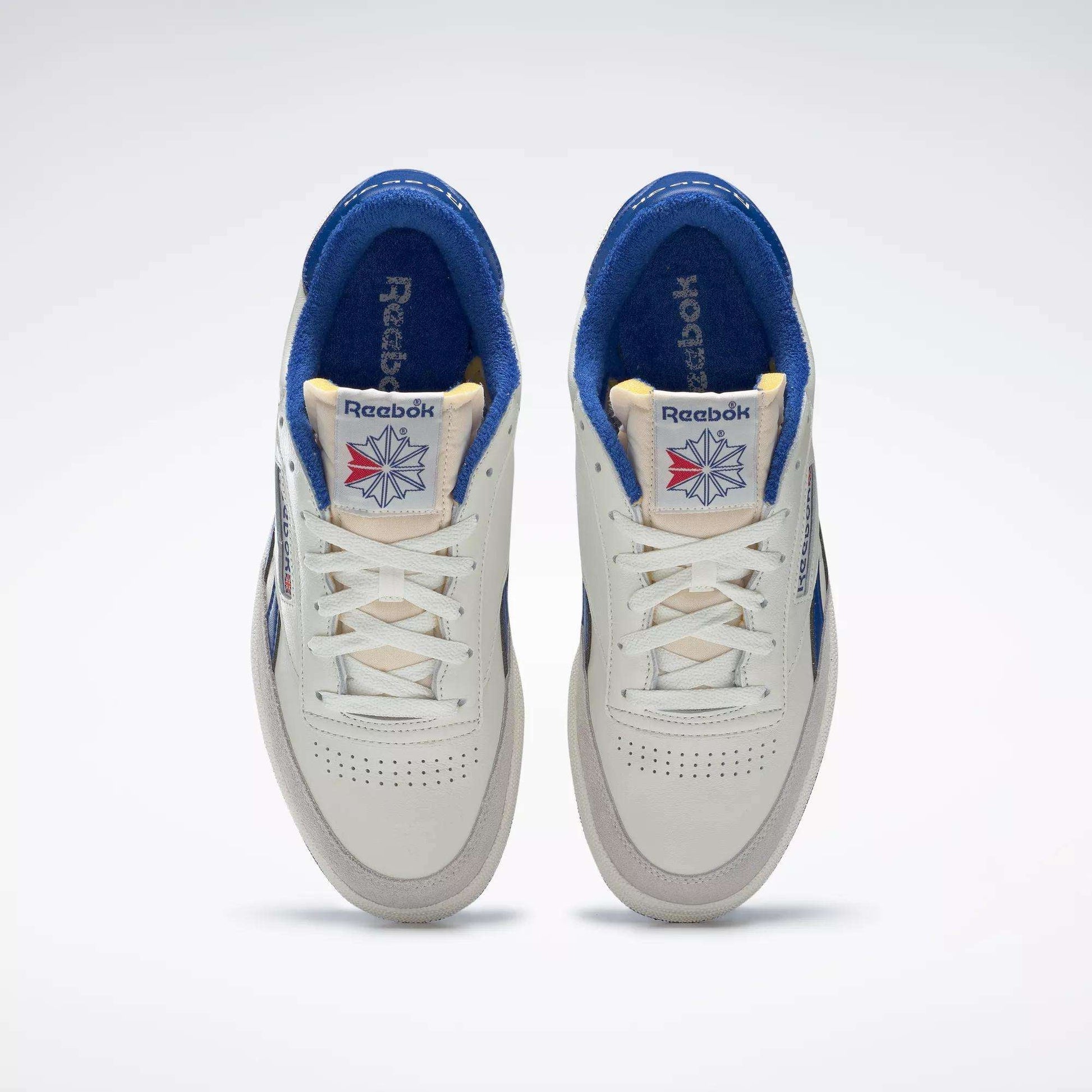 A pair of Reebok Men's Club C Revenge Vintage Shoes in white with blue accents, seen from above. These tennis-inspired sneakers have cream tongues, perforated toes, and tongue labels featuring the Reebok logo with a red and blue starburst.