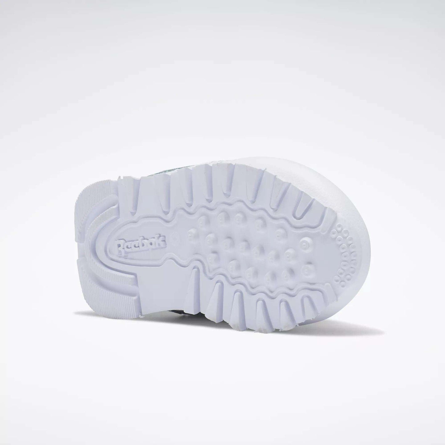 The image shows the white rubber sole of Reebok Kids' Classic Leather Shoes for babies and toddlers, viewed from below. The textured pattern provides grip, and the Reebok logo appears in raised letters near the heel.