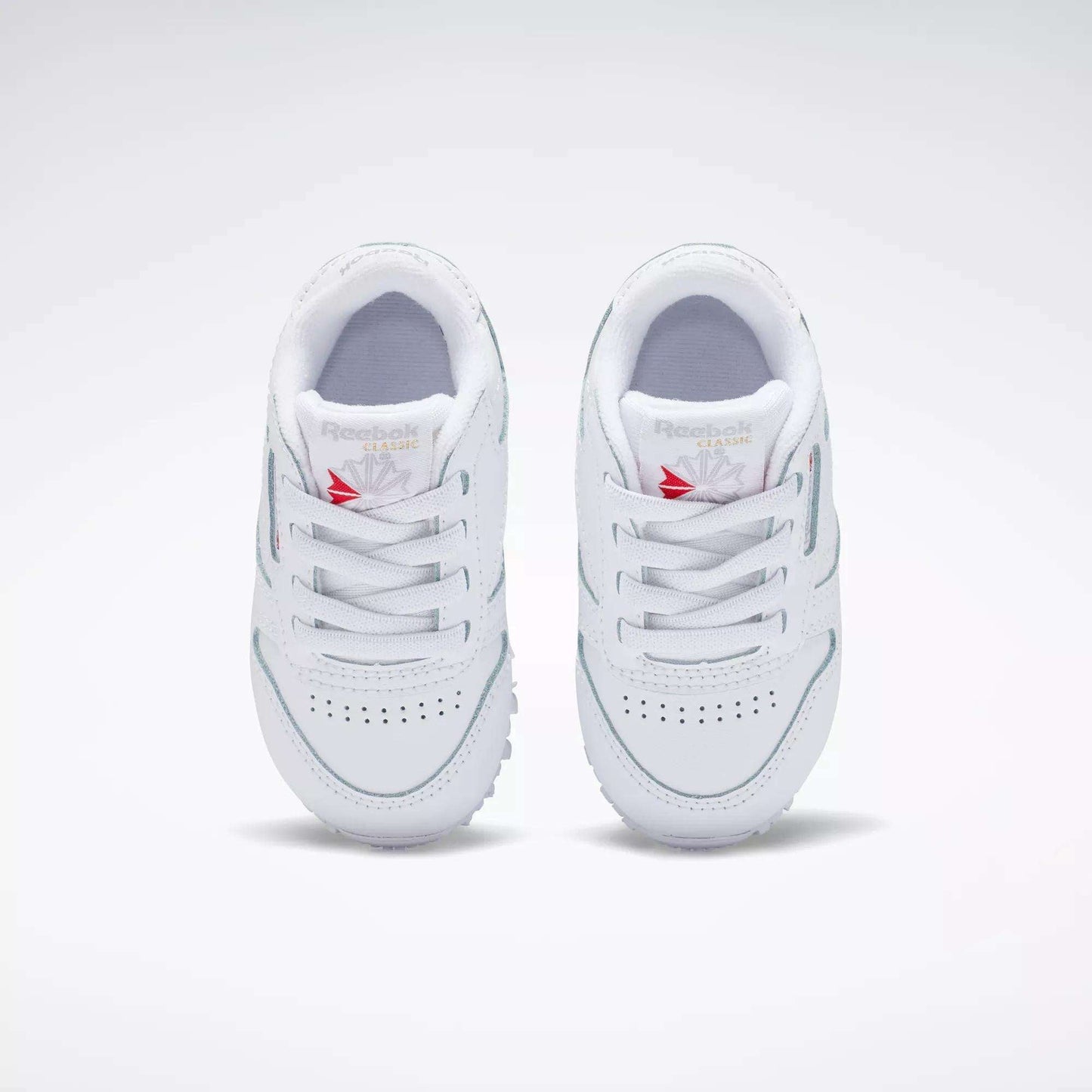 A pair of Kids' Classic Leather Shoes - Baby & Toddler by Reebok in white, featuring laces, toe perforations, and a red Reebok logo on the tongue, shown from above on a plain light background—ideal for toddlers’ everyday style.