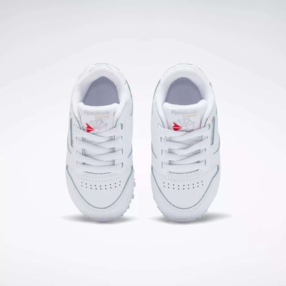 A pair of Kids' Classic Leather Shoes - Baby & Toddler by Reebok in white, featuring laces, toe perforations, and a red Reebok logo on the tongue, shown from above on a plain light background—ideal for toddlers’ everyday style.