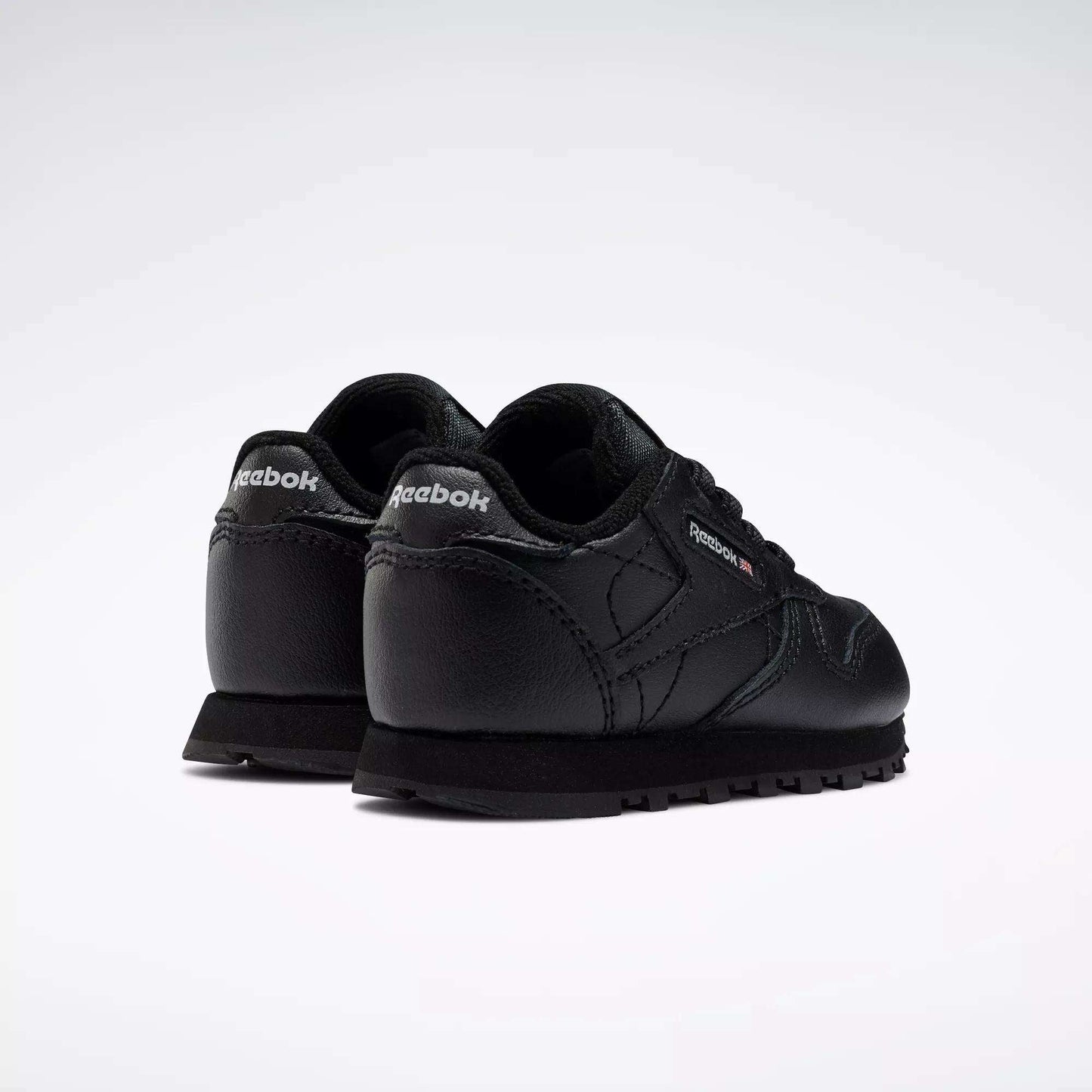 A pair of Kids' Classic Leather Shoes - Baby & Toddler by Reebok in black, shown from the back and side with visible Reebok logos on the heel and outer side, classic old-school style on a plain white background.