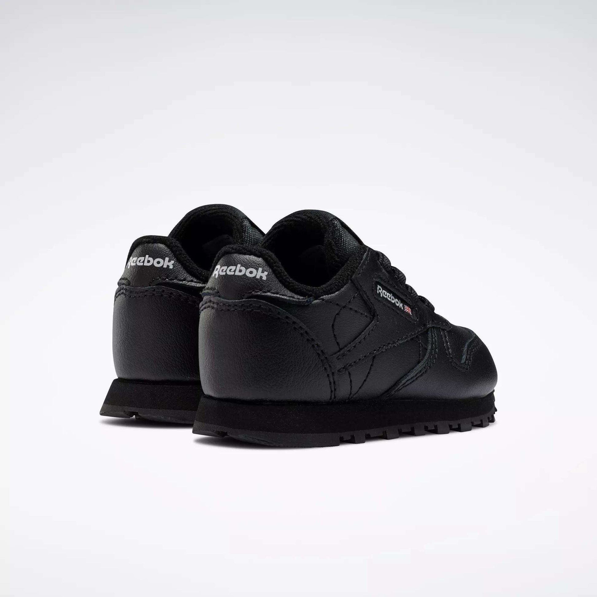 A pair of Kids' Classic Leather Shoes - Baby & Toddler by Reebok in black, shown from the back and side with visible Reebok logos on the heel and outer side, classic old-school style on a plain white background.