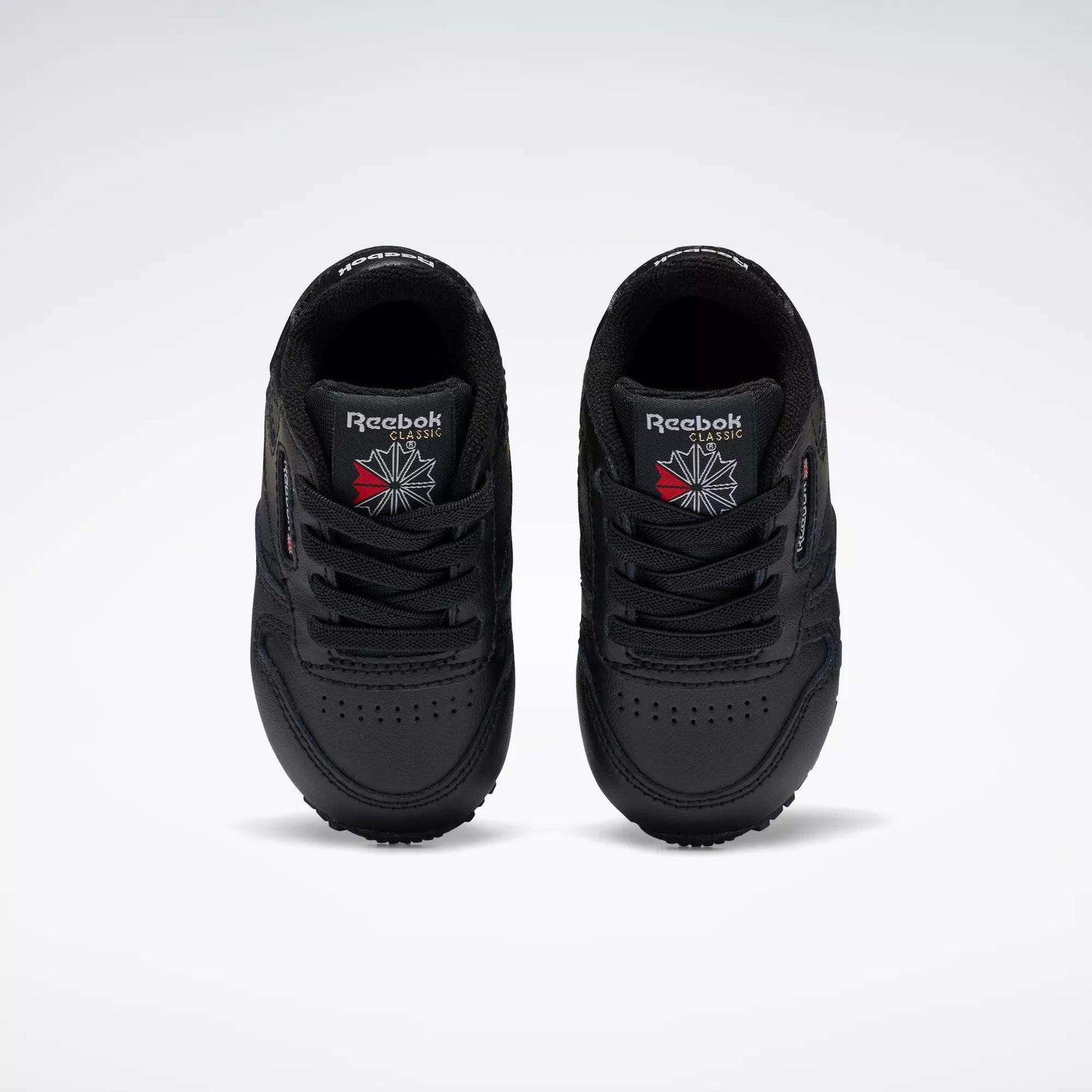 A pair of Reebok Kids' Classic Leather Shoes for baby & toddler in black, viewed from above with the Reebok logo and a red and white emblem on the tongues, set against a plain white background—ideal for young fans of classic sneakers.