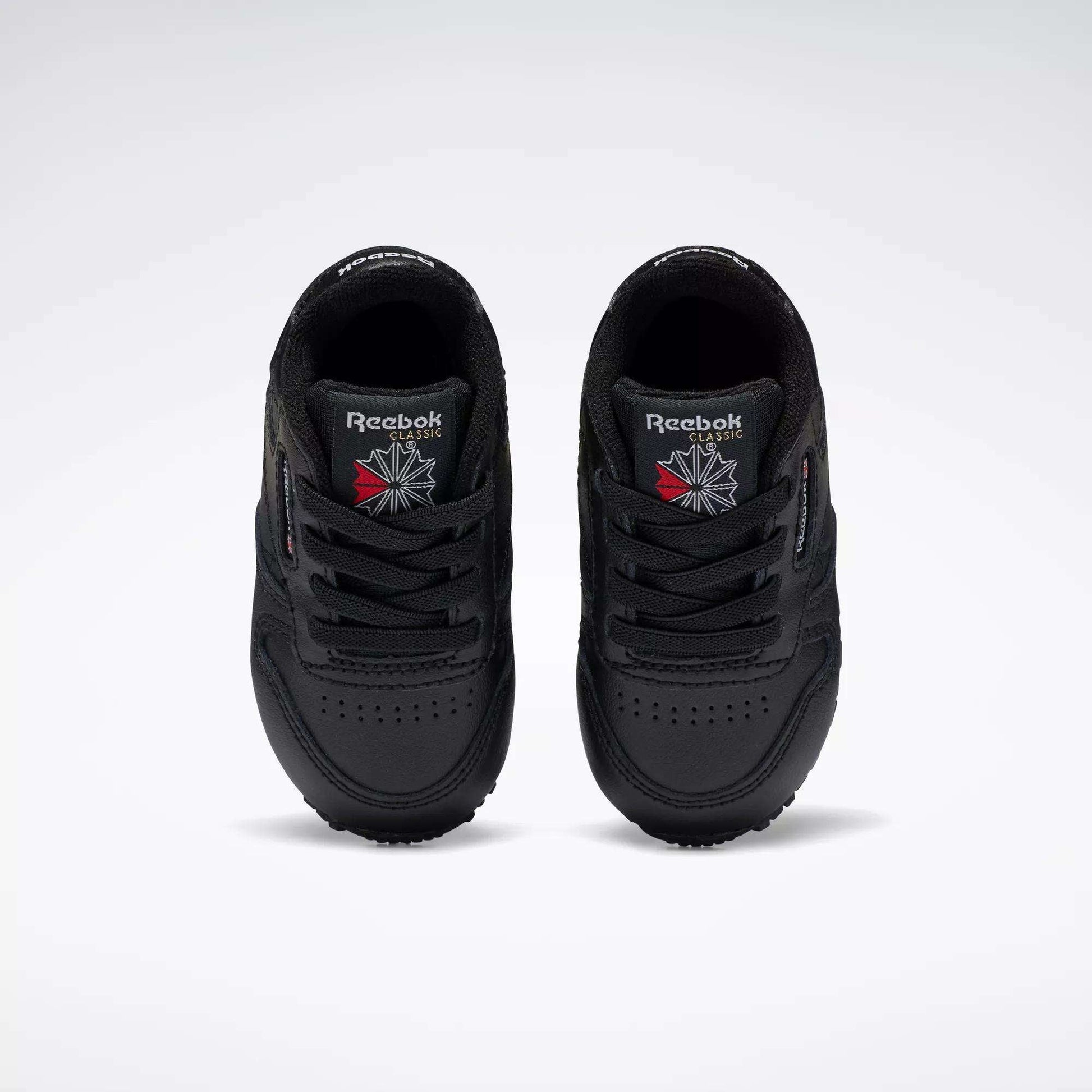 A pair of Reebok Kids' Classic Leather Shoes for baby & toddler in black, viewed from above with the Reebok logo and a red and white emblem on the tongues, set against a plain white background—ideal for young fans of classic sneakers.