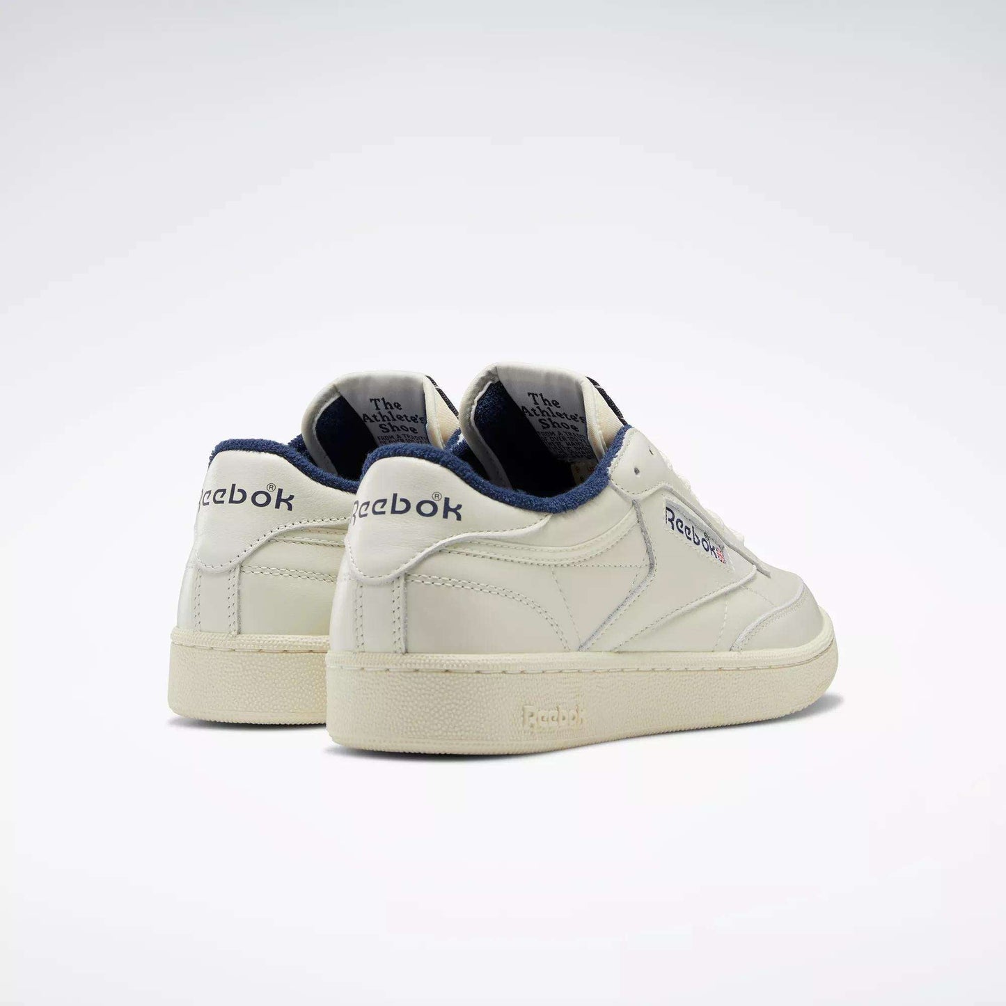 A pair of Reebok Club C 85 Vintage shoes in off-white with navy accents, featuring the Reebok logo on the side and heel. The retro design has a leather upper and beige soles, shown against a plain white background.