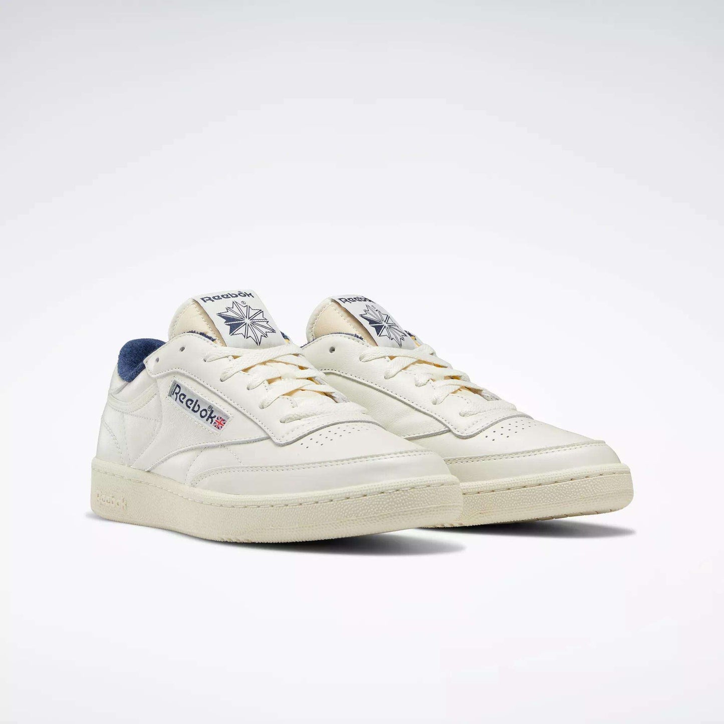 A pair of off-white Reebok Club C 85 Vintage Shoes with navy accents, featuring the Reebok logo and Union Jack on the side and tongue, along with lightweight cushioning for all-day comfort, set against a plain white background.