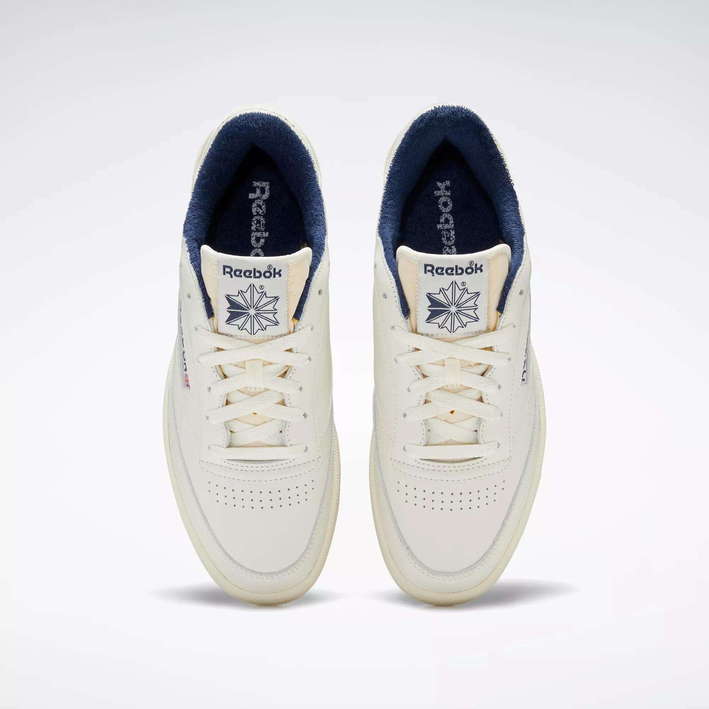 Top view of a pair of Reebok Club C 85 Vintage Shoes in off-white with a leather upper, navy blue inner lining, perforated toe boxes, and Reebok logo with star on the tongues.
