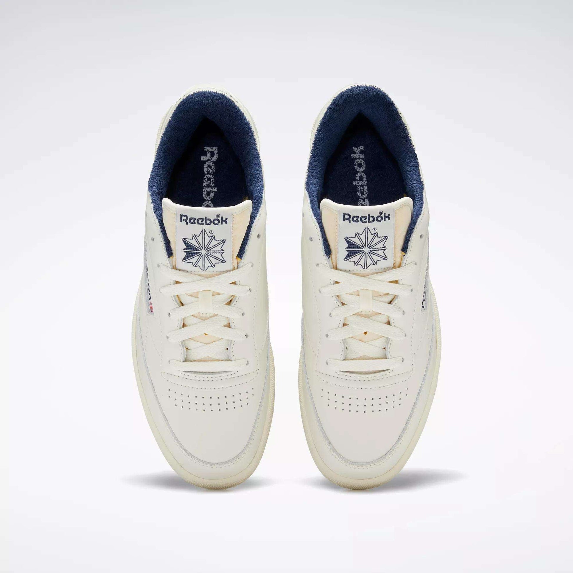 Top view of a pair of Reebok Club C 85 Vintage Shoes in off-white with a leather upper, navy blue inner lining, perforated toe boxes, and Reebok logo with star on the tongues.