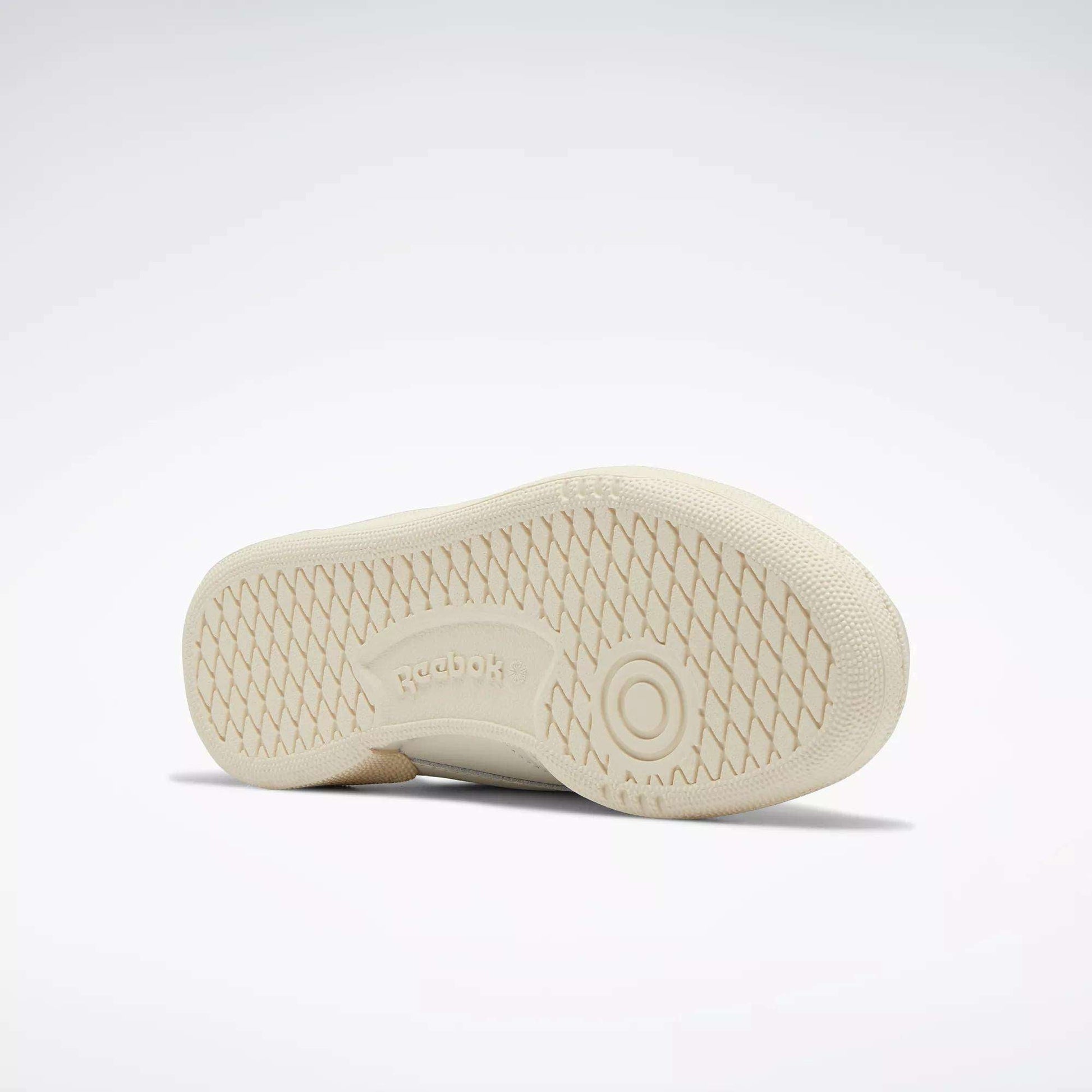 A Women's Club C 85 Vintage Shoe by Reebok in beige rests on its side, revealing a grid-patterned rubber sole with the Reebok logo at the center. The background is plain and light-colored.