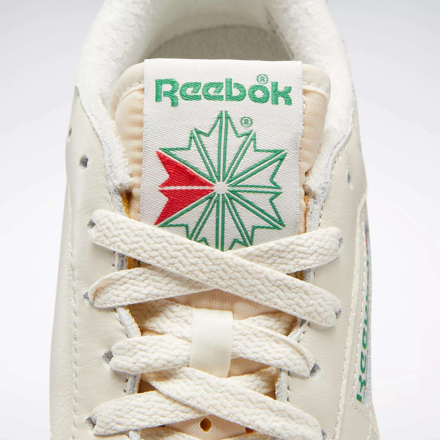 Close-up of Reebok Women's Club C 85 Vintage Shoes, featuring the green Reebok logo above a green and red starburst on the tongue, with cream laces and textured leather for a stylish, classic tennis look.