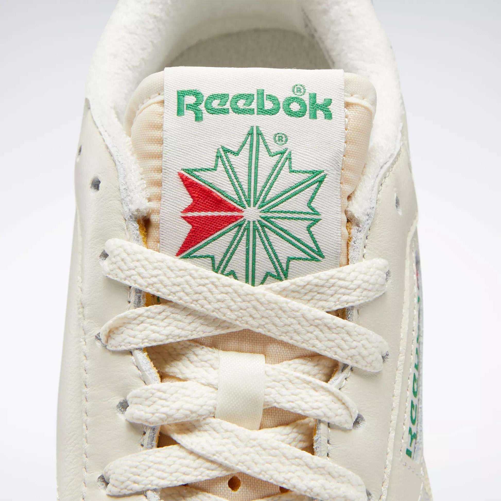 Close-up of Reebok Women's Club C 85 Vintage Shoes, featuring the green Reebok logo above a green and red starburst on the tongue, with cream laces and textured leather for a stylish, classic tennis look.