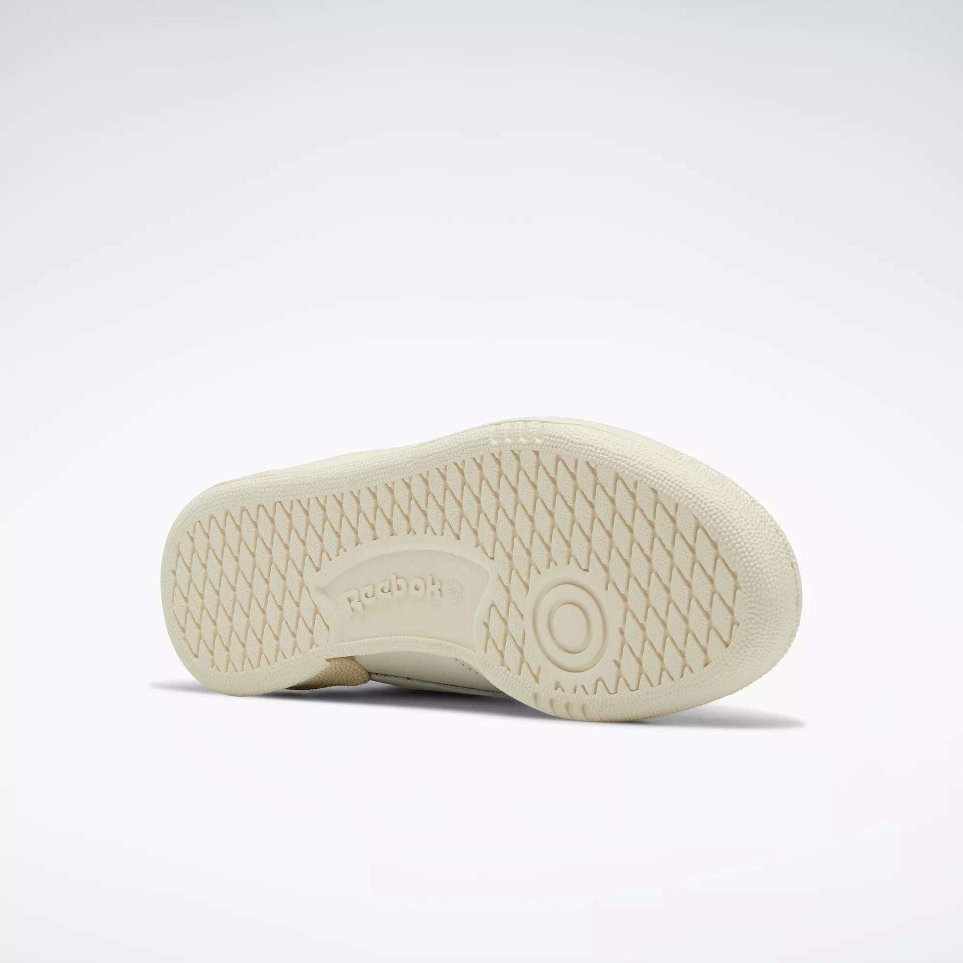 A Women's Club C 85 Vintage Shoe by Reebok rests on its side, showcasing a textured sole with a circular grip and an embossed Reebok logo at the center. This tennis-inspired sneaker pops against a light gray background.