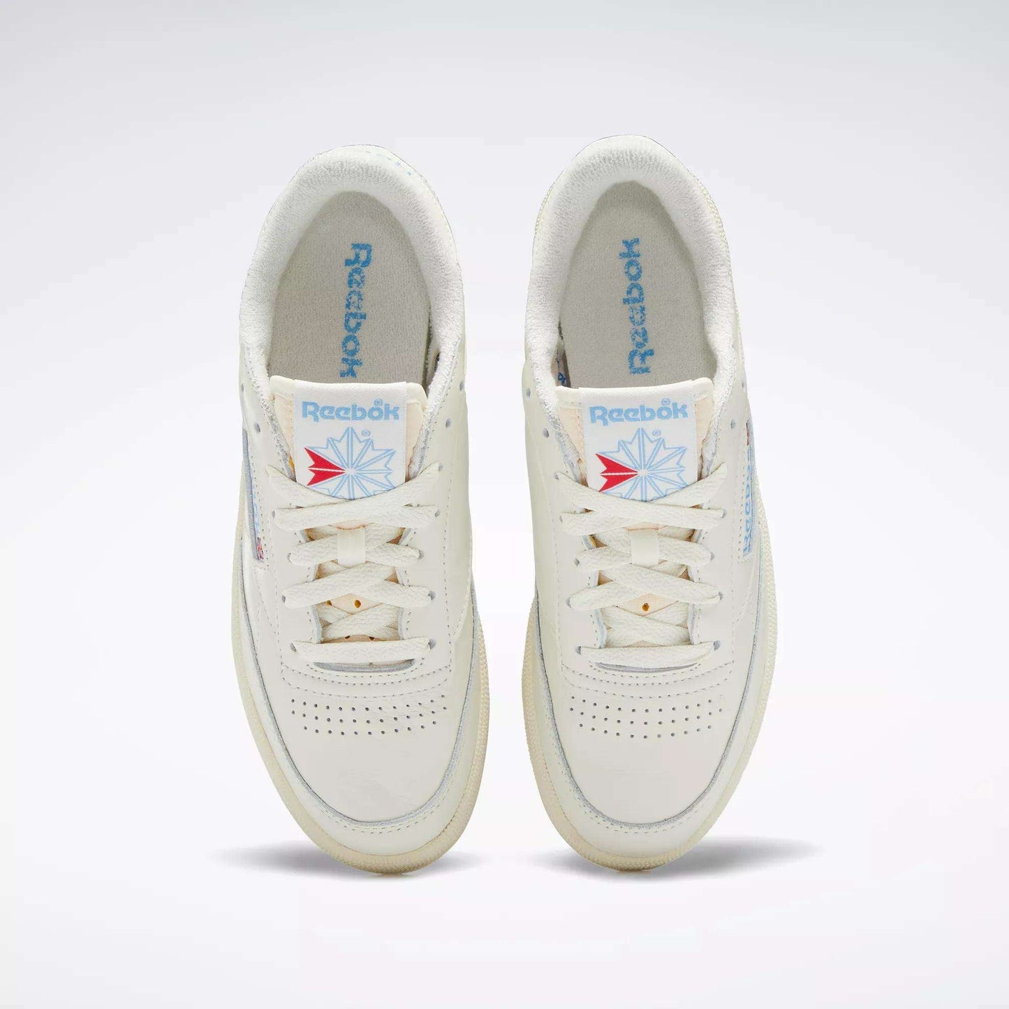 A pair of Reebok Women's Club C 85 Vintage Shoes in off-white, seen from above, with blue Reebok logos on the tongues and insoles, white laces, and subtle perforations on the toe boxes.