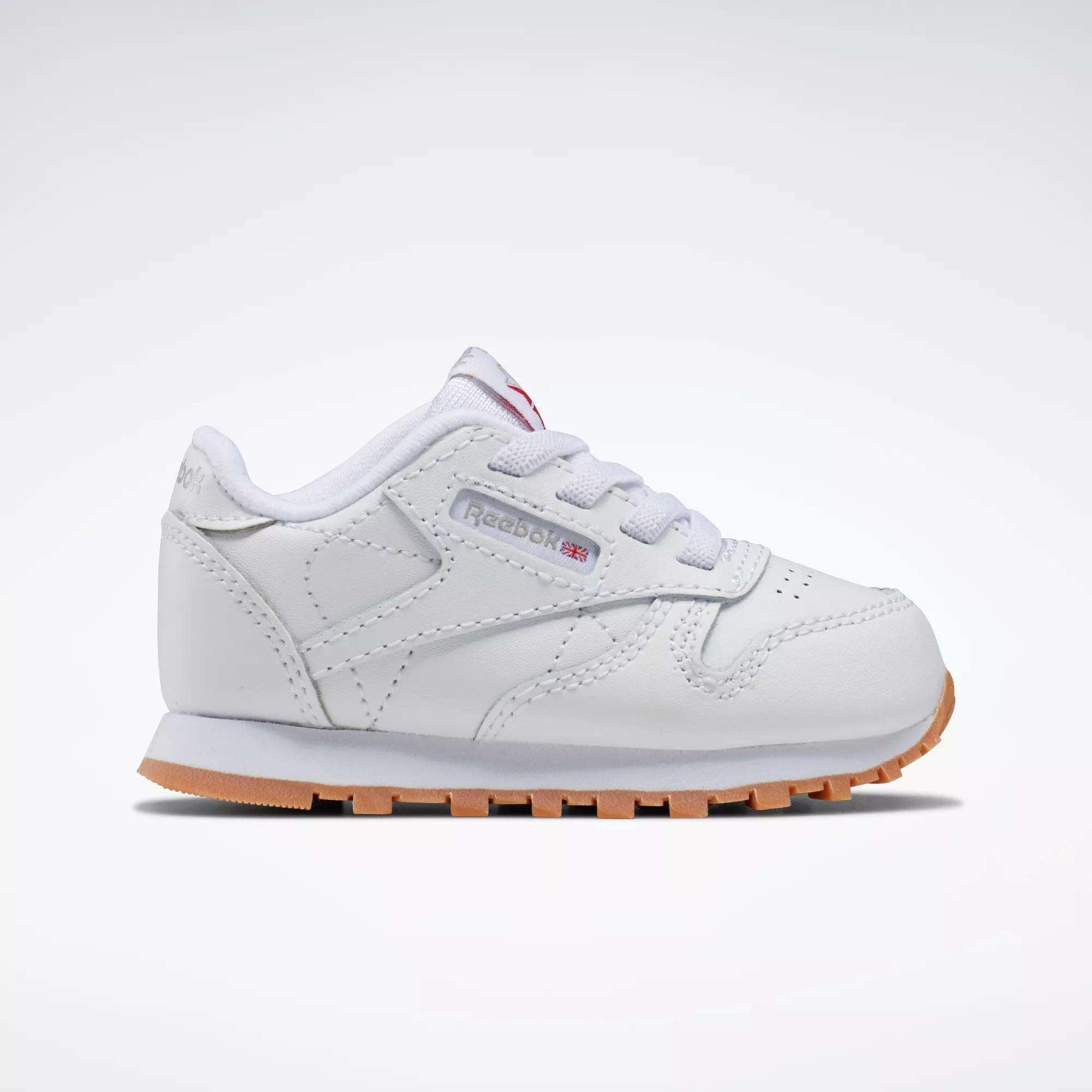 The Reebok Kids' Classic Leather Shoes for babies and toddlers are white with a light brown sole, white laces, and a side tag featuring the Reebok logo, shown from the side on a plain white background.