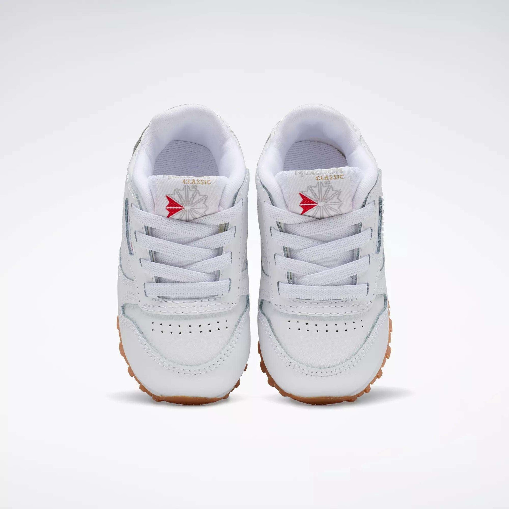 The Reebok Kids' Classic Leather Shoes - Baby & Toddler feature white leather, gum-colored soles, white laces, and a red and gray starburst logo on the tongue—perfect for active toddlers.