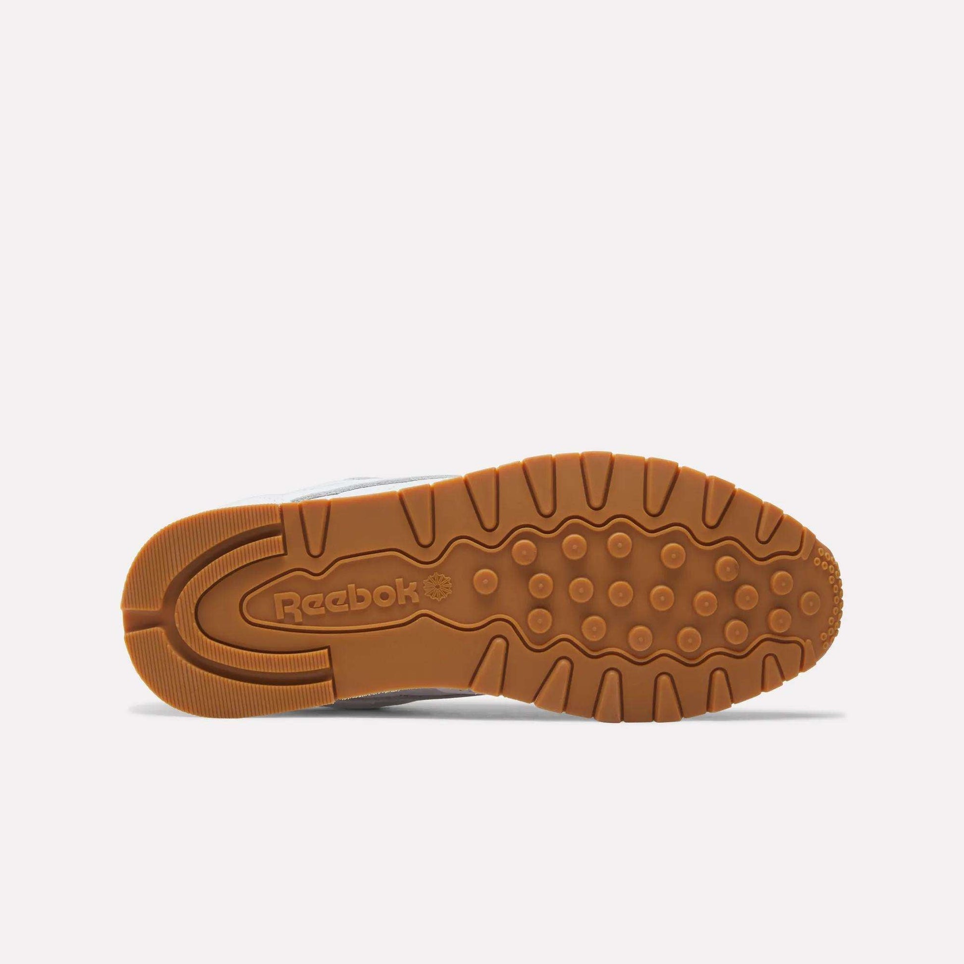 The image displays the bottom view of Reebok Classic Leather Shoes with a brown rubber sole, textured grip patterns, and the Reebok logo embossed near the heel, showcasing its retro-inspired design.