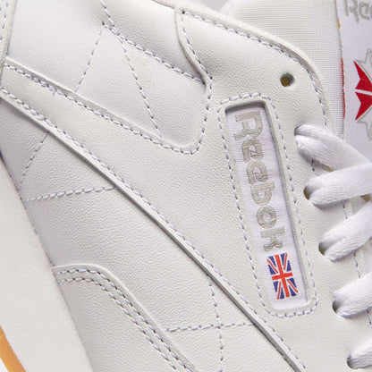 Close-up of Reebok Classic Leather Shoes featuring durable white leather, embossed branding, a small Union Jack flag on the side, and visible white stitching and laces for a timeless retro look.