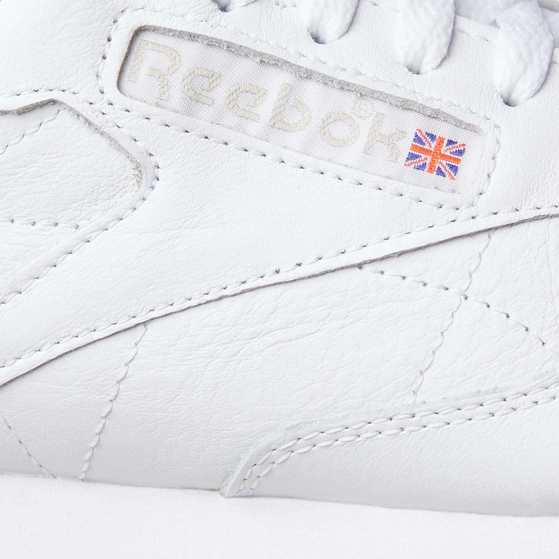 Close-up of Reebok Classic Leather Shoes in white, spotlighting the stitched Reebok logo with a Union Jack flag on the side. Features include a durable leather upper and retro-inspired stitching details.