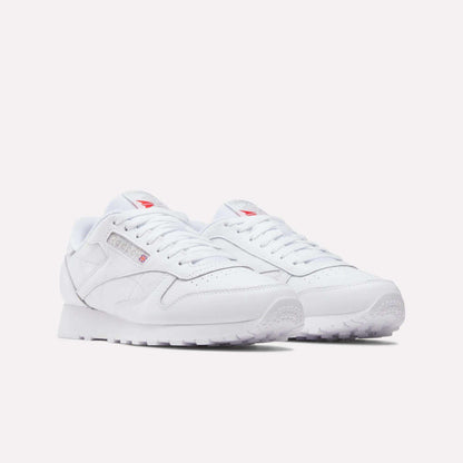 The Reebok Classic Leather Shoes feature a minimal design with a leather upper, white laces, and subtle red Reebok branding on the tongue and side, shown against a plain light background.