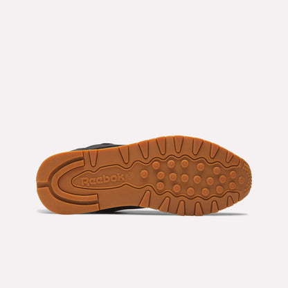 The image displays the bottom view of Reebok’s Classic Leather Shoes, showcasing a brown gum rubber sole with the Reebok logo and textured patterns for traction, emphasizing the retro style of these sneakers.
