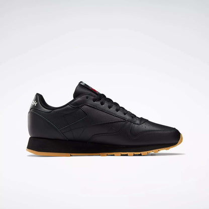 Side view of Reebok Classic Leather Shoes in black with a gum sole, black laces, minimal stitching, and low-cut design. These retro-inspired shoes have a durable leather upper and are shown on a plain light gray background.