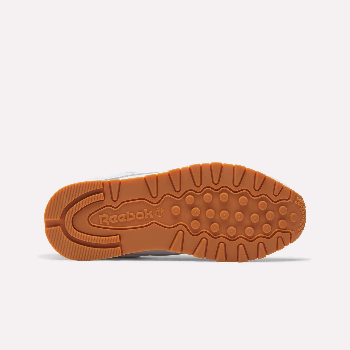 The image displays the sole of Reebok Women's Classic Leather Shoes with a light brown rubber outsole, textured patterns for grip, and an engraved Reebok logo near the heel. The cushioned midsole offers comfort. Background is plain white.