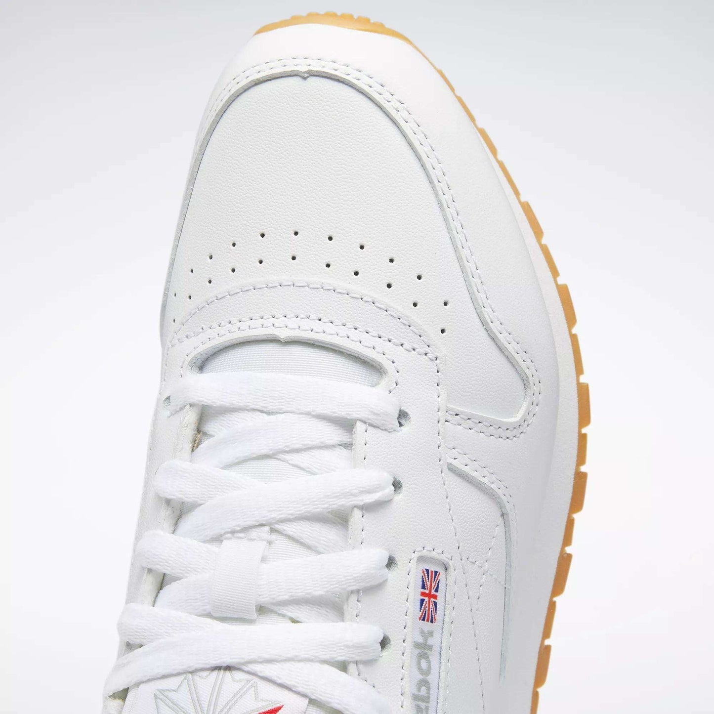 Close-up of Reebok Women's Classic Leather Shoes in white, featuring white laces, perforated toe, cushioned midsole, small British flag logo on the side, and a light brown rubber sole.
