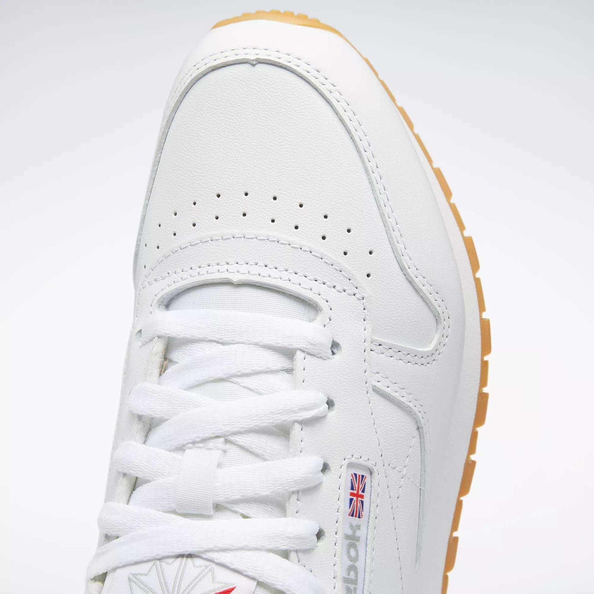 Close-up of Reebok Women's Classic Leather Shoes in white, featuring white laces, perforated toe, cushioned midsole, small British flag logo on the side, and a light brown rubber sole.