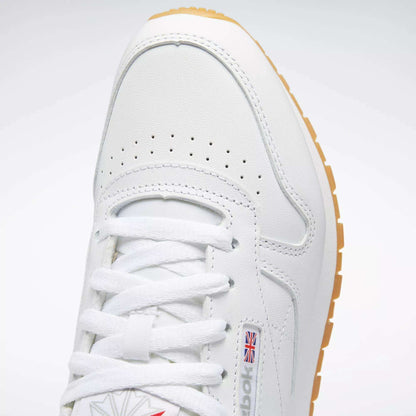 Close-up of Reebok Women's Classic Leather Shoes in white, featuring white laces, perforated toe, cushioned midsole, small British flag logo on the side, and a light brown rubber sole.