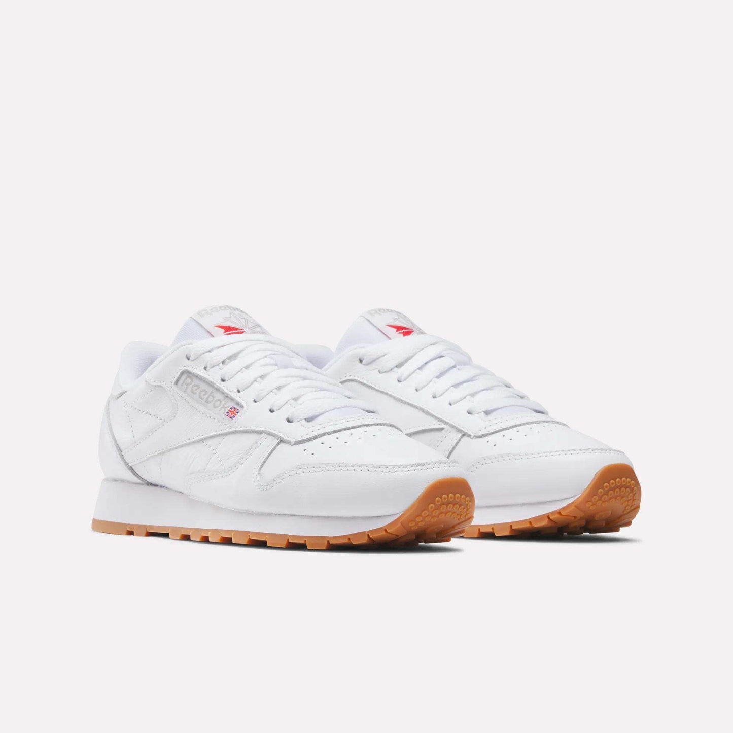 A pair of Reebok Women's Classic Leather Shoes with gum rubber soles is displayed on a light background. These leather shoes feature white laces, subtle branding, a low-cut design, and a cushioned midsole for comfort.
