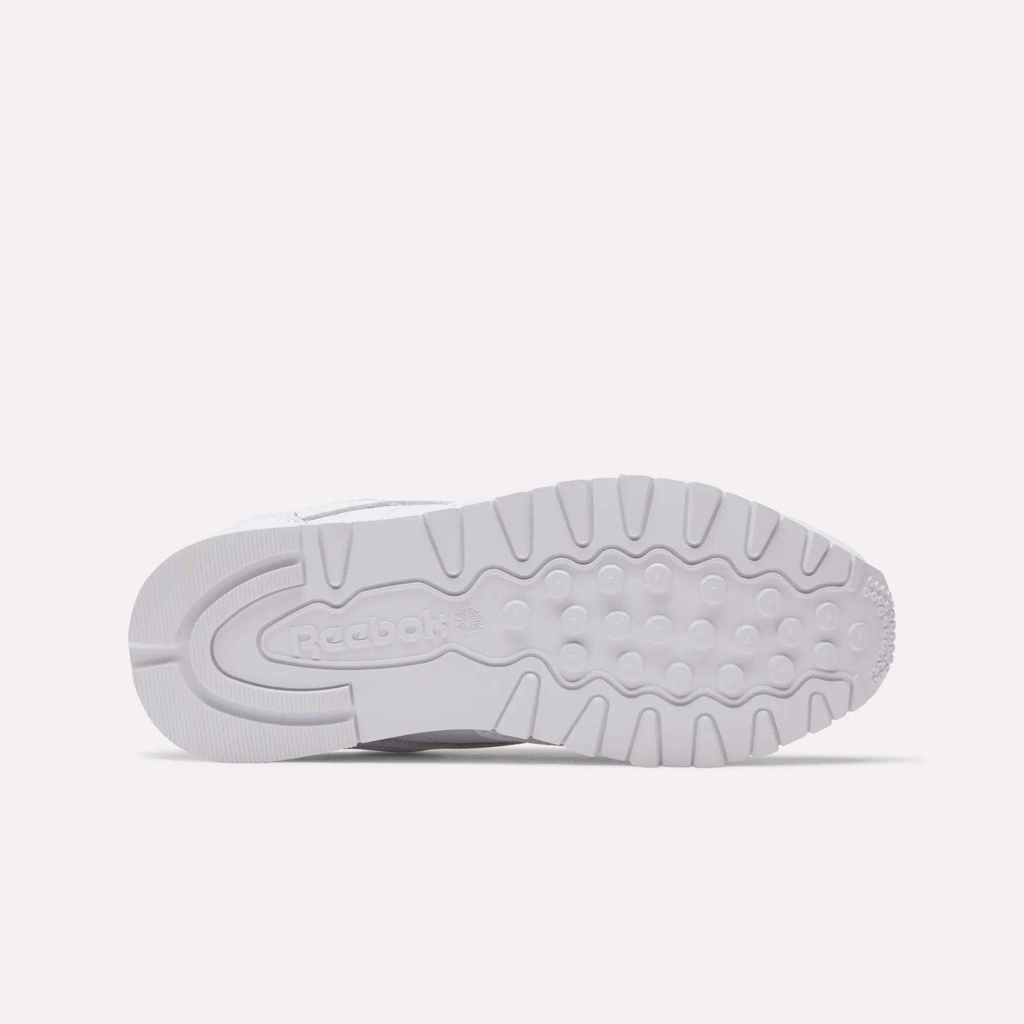 The image displays the outsole of a Women's Classic Leather Shoe by Reebok, featuring white rubber with grooves, circular treads, and an embossed Reebok logo near the heel. The sneaker is shown lying on its side.