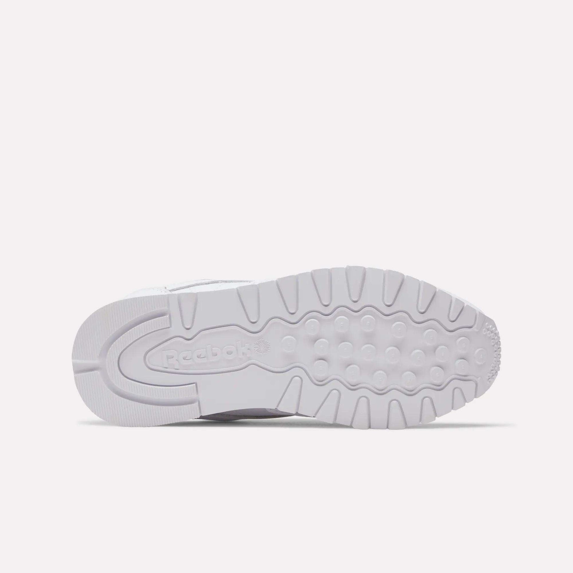 The image displays the outsole of a Women's Classic Leather Shoe by Reebok, featuring white rubber with grooves, circular treads, and an embossed Reebok logo near the heel. The sneaker is shown lying on its side.