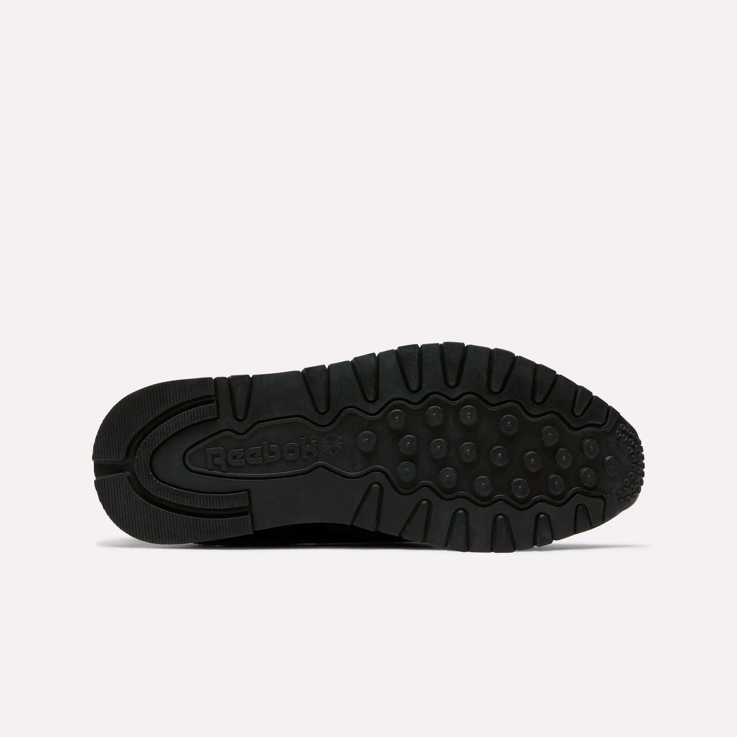 The image shows the black rubber sole of the Reebok Women's Classic Leather Shoes, viewed from below. It features circular tread patterns, grooves for traction, Reebok branding near the heel, and a cushioned midsole for comfort.
