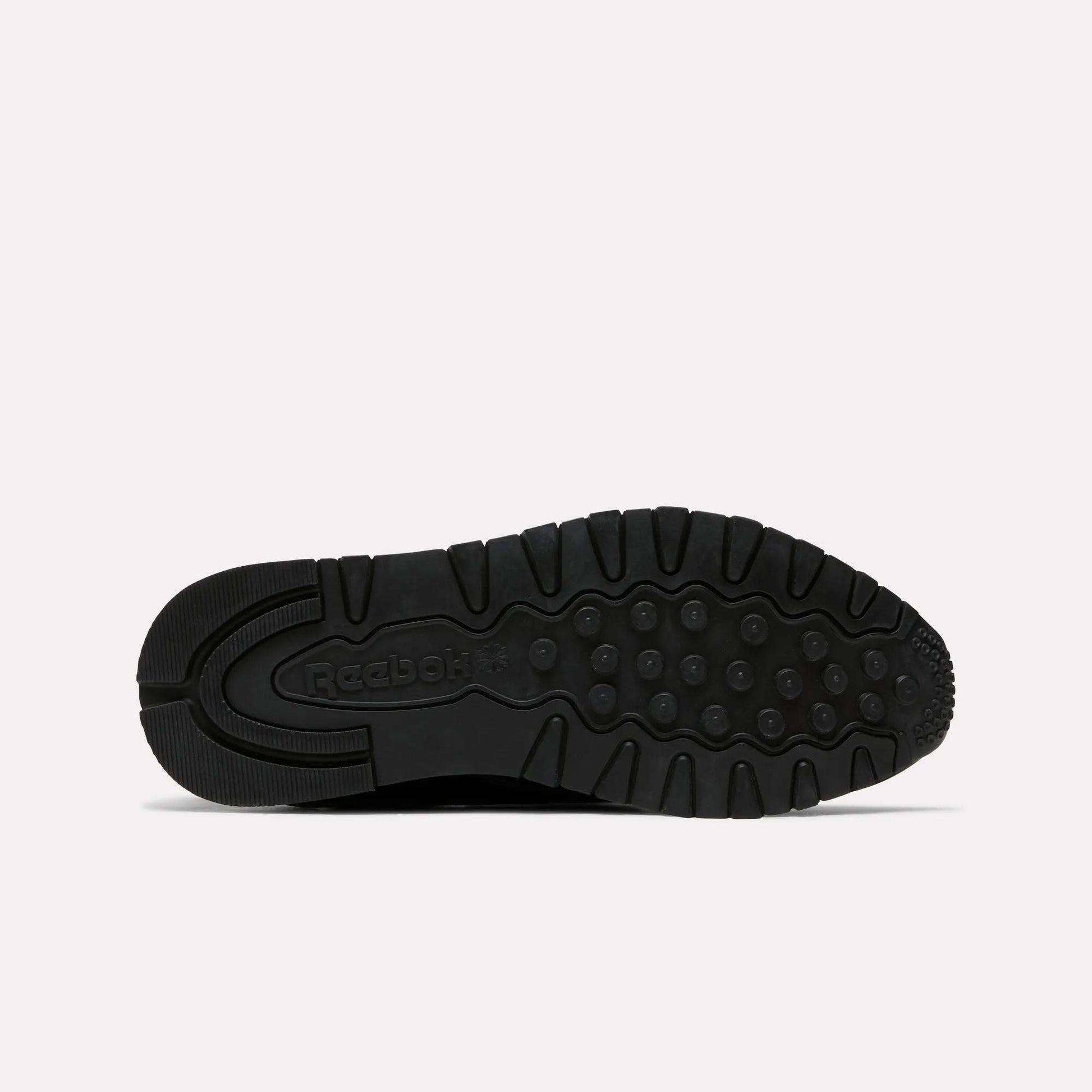 The image shows the black rubber sole of the Reebok Women's Classic Leather Shoes, viewed from below. It features circular tread patterns, grooves for traction, Reebok branding near the heel, and a cushioned midsole for comfort.