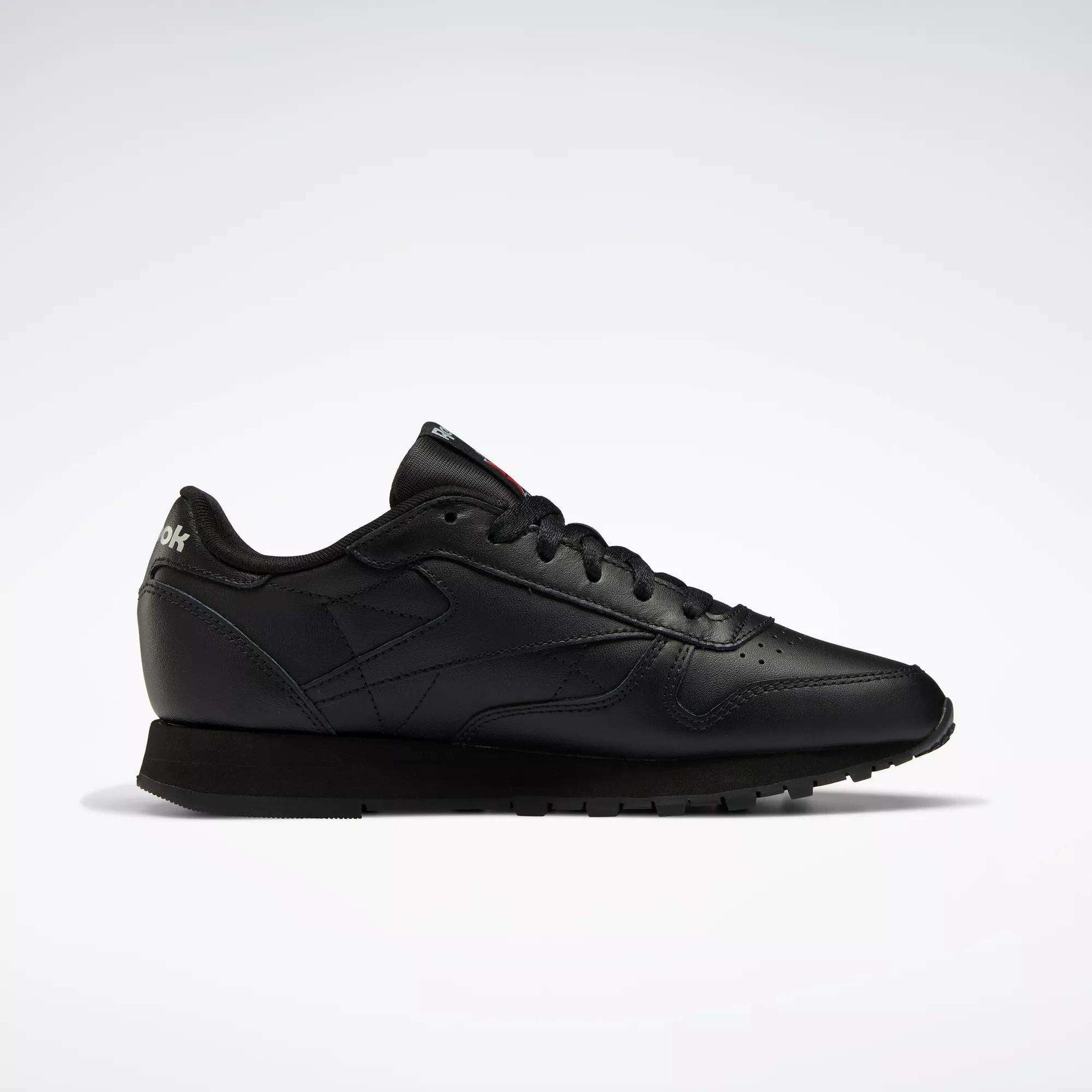 The Reebok Women's Classic Leather Shoes are shown in profile on a white background, featuring a low-top black design, cushioned midsole, black laces, and a textured rubber sole.