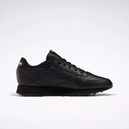 The Reebok Women's Classic Leather Shoes are shown in profile on a white background, featuring a low-top black design, cushioned midsole, black laces, and a textured rubber sole.