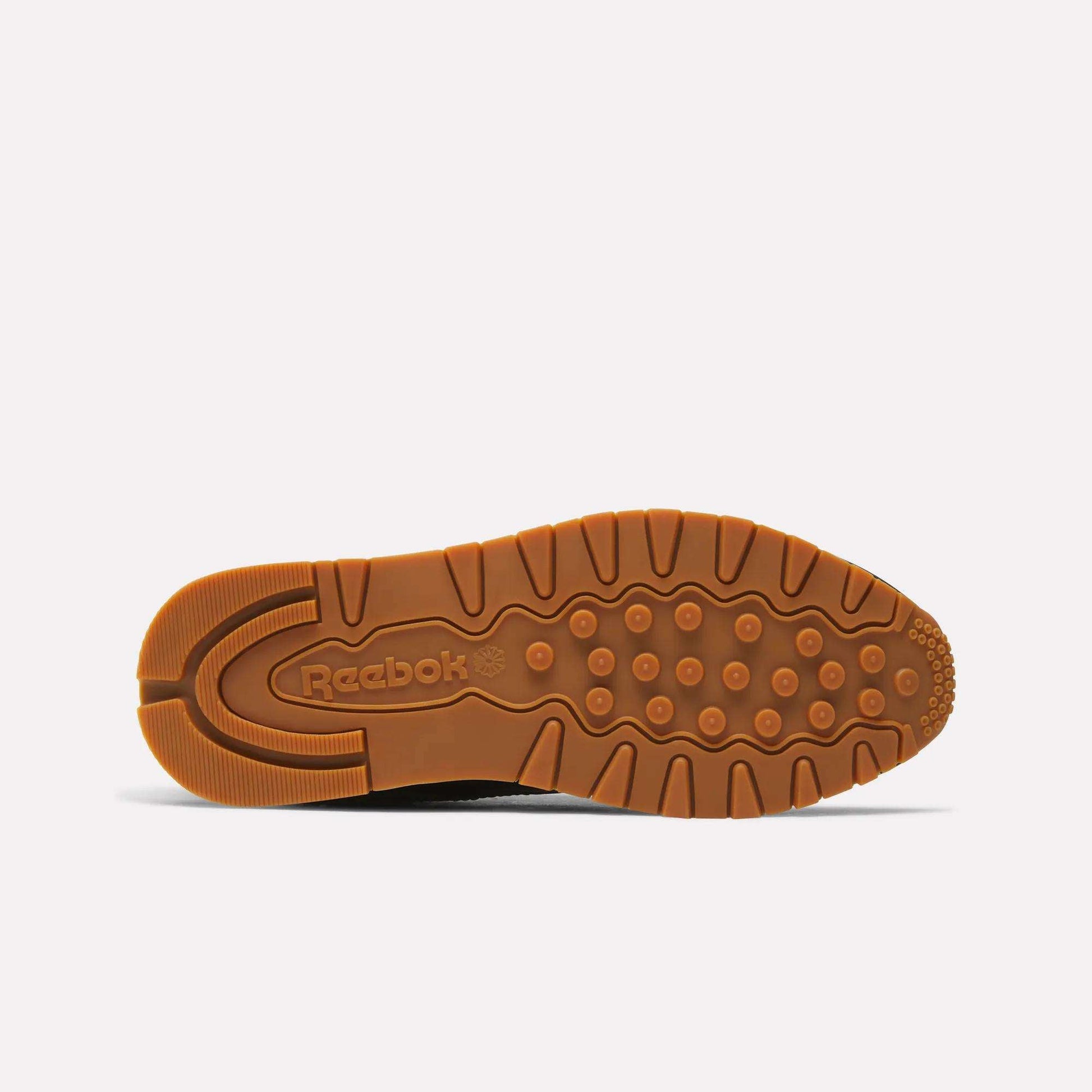 The image shows the sole of a Reebok Women's Classic Leather Shoe, featuring a brown rubber outsole with circular grips and an embossed Reebok logo by the heel. The cushioned midsole adds comfort, all set against a plain light background.