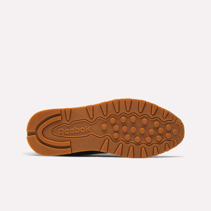 The image shows the sole of a Reebok Women's Classic Leather Shoe, featuring a brown rubber outsole with circular grips and an embossed Reebok logo by the heel. The cushioned midsole adds comfort, all set against a plain light background.