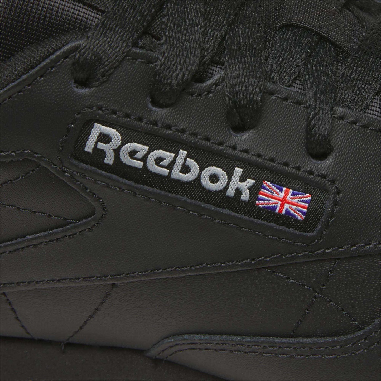 Close-up of Reebok Women's Classic Leather Shoes in black, showcasing the Reebok logo, a small Union Jack flag by the laces, and a cushioned midsole for comfort.
